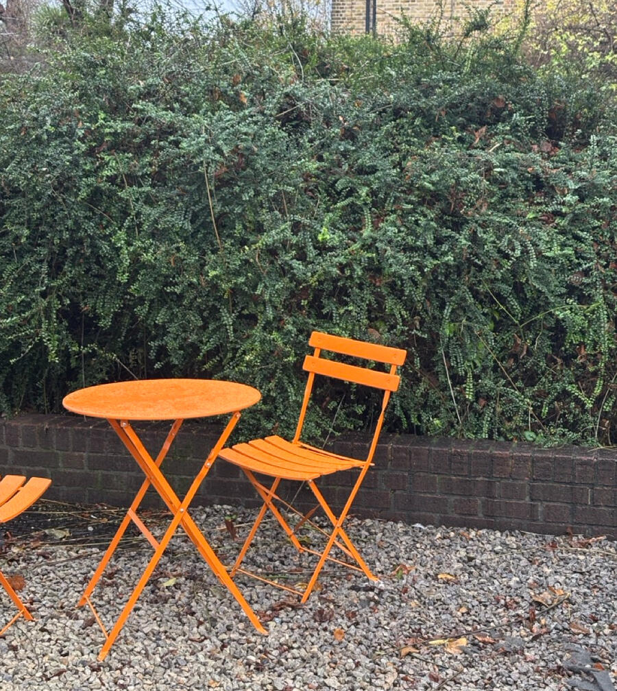A photo of a small, bright orange table with two chairs, situated outdoors on a gravel floor. Behind it is a somewhat overgrown dark green hedge.