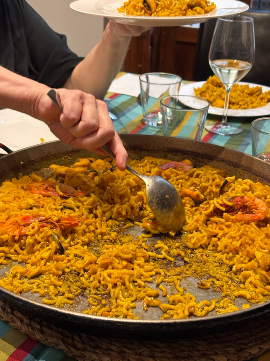 A paella dish covered in small yellow noodles and seafood. A hand is holding a spoon and scraping food out to a waiting plate.