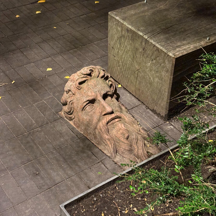 Stone sculpture of a bearded face lying on the ground beside a concrete bench and paving stones.