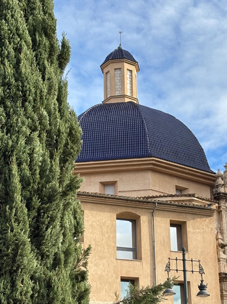 An upward angled view of a yellow Spanish-style building. The roof – the focal point of the photo – is a deep, dark blue that is vaguely dome-shaped. The left side of the shot shows greenery from a tree in the foreground, partially obstructing the view of
