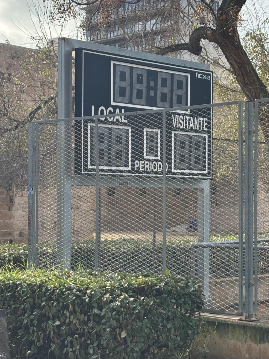 A digital scoreboard partially obstructed by a fence. It's black and the scores are blank. One side reads "Local" while the other reads "Visitante".