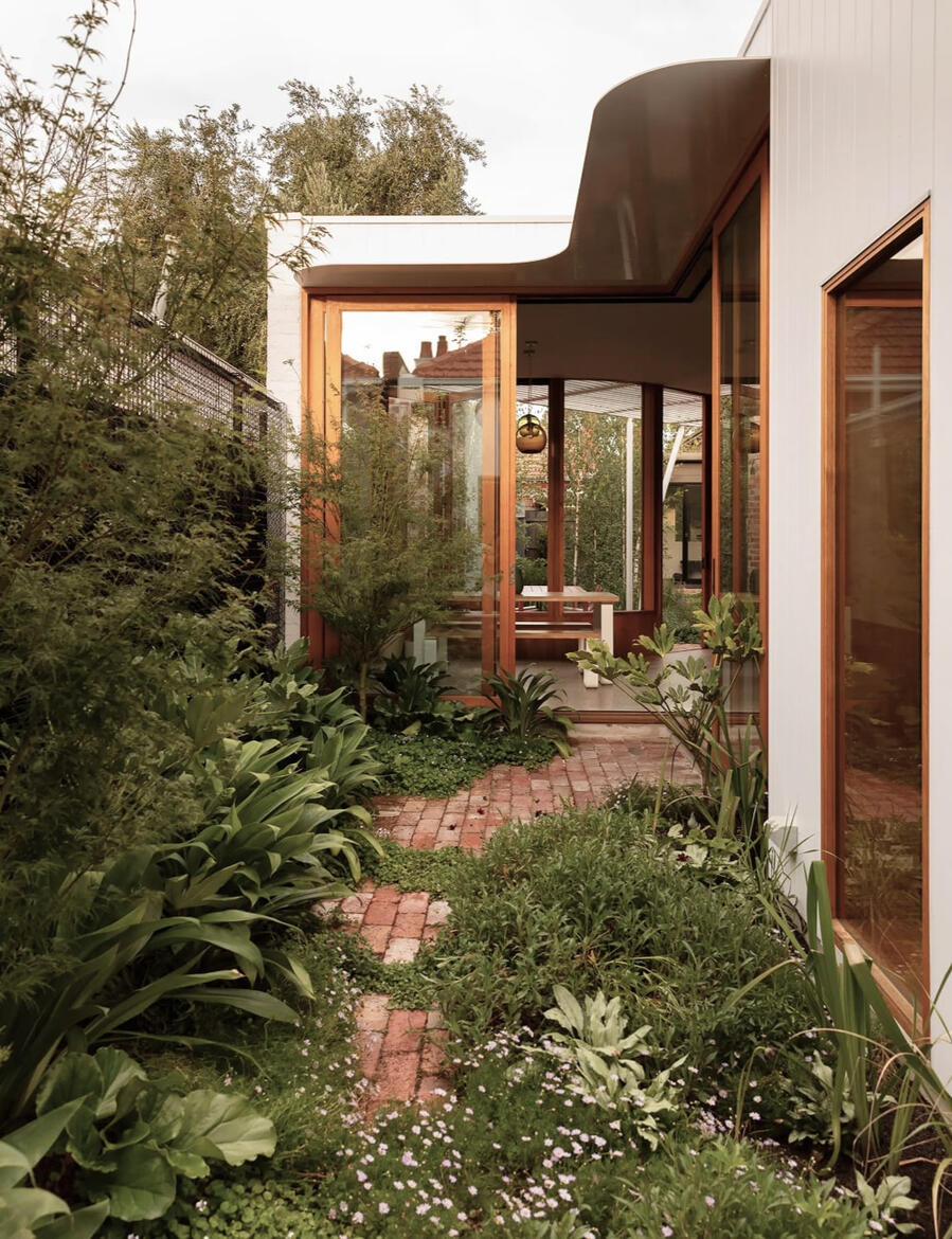 A garden path leading towards the glass-walled entrance of a modern-looking home. The red brick pathway is curved and charmingly overgrown with low-growing plants and bushes.
