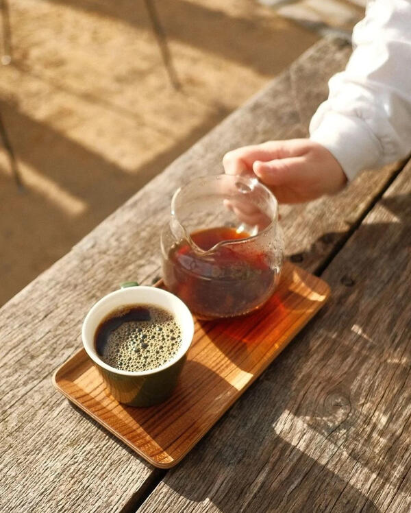 A cup of black filter coffee next to a small glass jug containing more, sitting on a wooden tray on a wooden table.