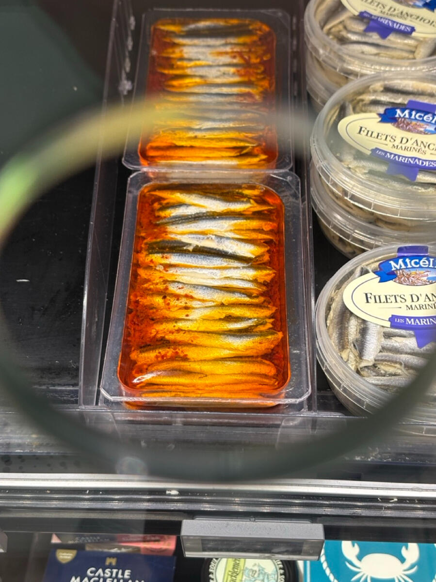 A container of anchovies submerged in a bright orange oil, sitting on a supermarket shelf next to other containers of fish.