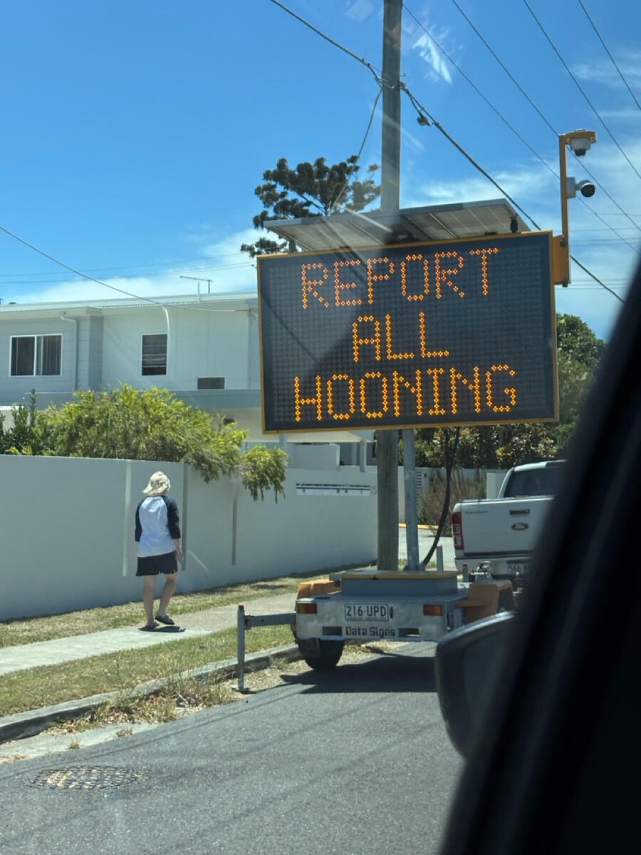 A side-of-the-road digital sign says "Report all hooning", against a blue-skied suburban backdrop.