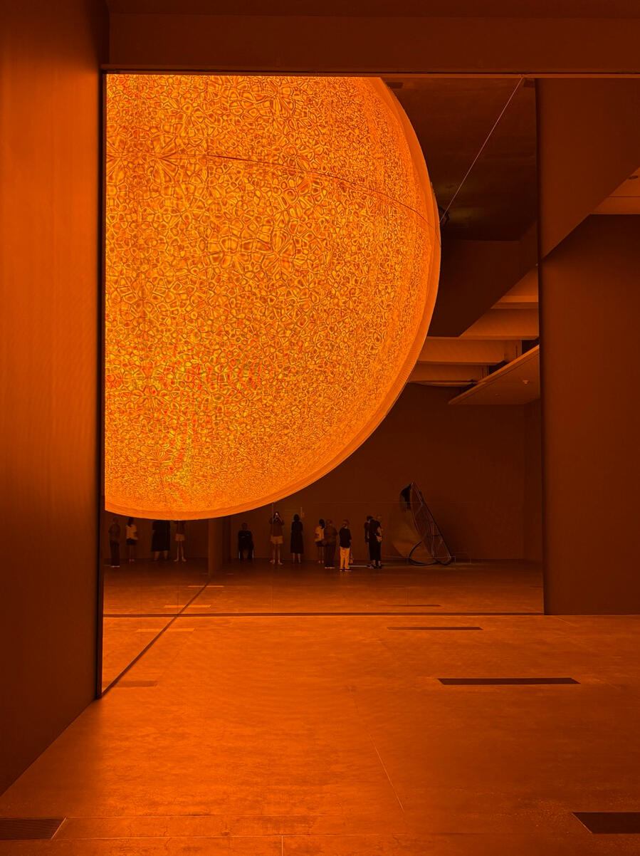 A large orange artificial sun hangs over a warehouse-like room of onlookers, who appear tiny in the distance.