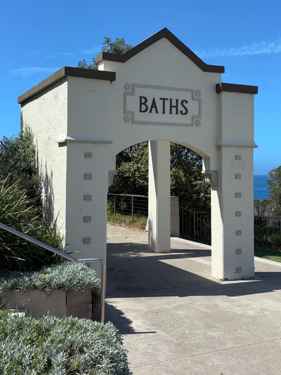 A sunlit archway sits over a plant-lined path. On the archway is retro lettering saying "Baths".
