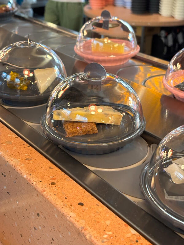 Small plates of cheese, covered by glass domes, sit on a tabletop conveyor belt.