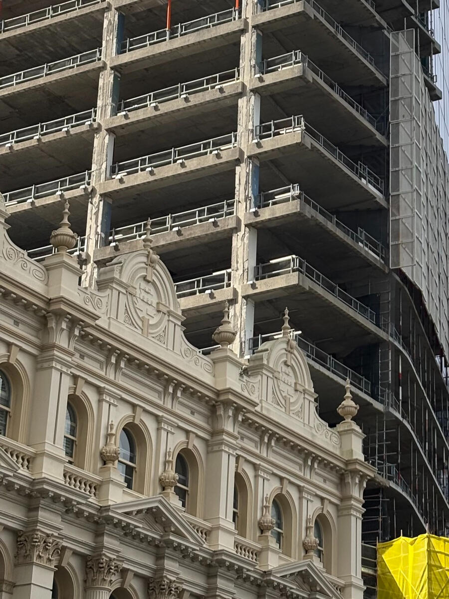A contrasting photo with a smaller classic building in the foreground, dwarfed by a much larger modern skyscraper in the background, still under construction.
