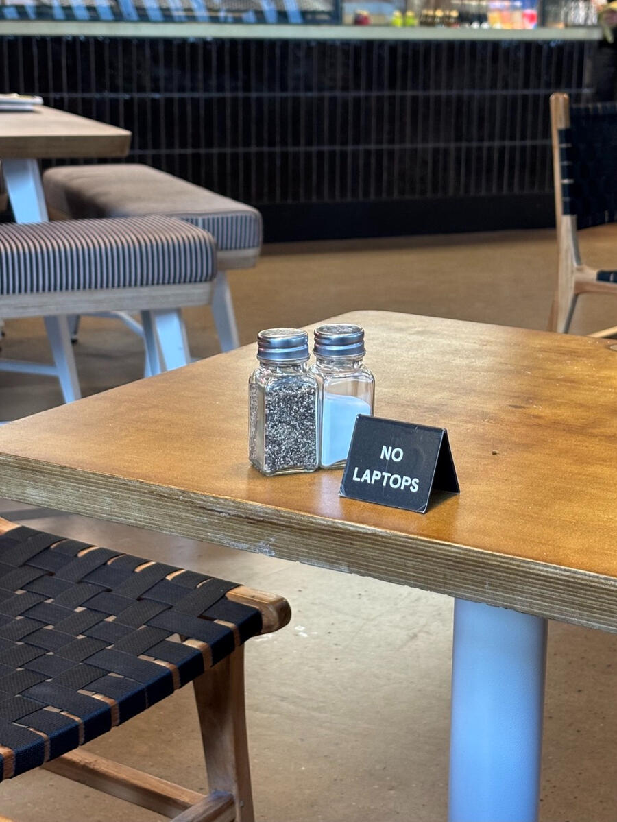 A salt shaker and pepper shaker sit on a wooden table. A small sign next to them says "No Laptops".