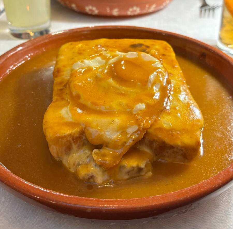 A sandwich sitting in a bowl, covered it a light brown sauce that is filling up the sides of the bowl. A fried egg and melted cheese are visible under the sauce, on top of the bread.