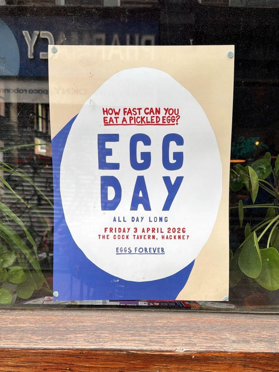 A paper sign is stuck up in a window. It says "How fast can you eat a pickled egg?", followed by a large title "EGG DAY", with small text below saying "All day long. Friday 3 April 2026. The Cock Tavern, Hackney. Eggs forever."